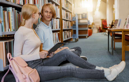 Friends student sit on floor near bookshelves in library and studying. Prepearing for examの写真素材