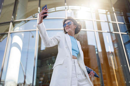 Pretty woman in trendy outfit making selfie on background of modern building