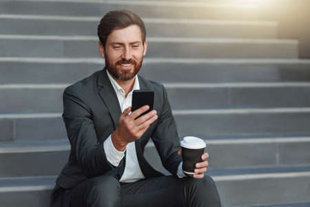 Smiling office worker in suit sitting on stairs with coffee during break and using phoneの写真素材