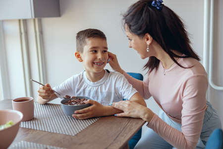 Portrait, small teen boy eating cereals with milk in kichen and mother touching his cheek.の写真素材