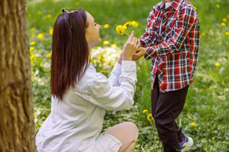 Beautiful young mother getting flowers from small son in park. Woman with dandelions from kid.