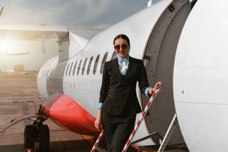 Woman flight attendant in sunglasses standing on airplane stairs at airport and looking camera