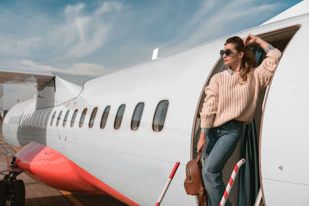 Woman passenger in sunglasses standing on airplane stairs at airport and looking awayの写真素材