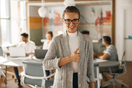 Portrait of smiling businesswoman in suit standing in modern office and showing thumb up