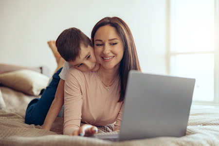 Happy beautiful young Caucasian woman lying on bed with laptop, small son hugging behind.の写真素材