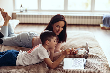 Small Caucasian cute boy with beautiful mother lying together on bed with laptop.の写真素材