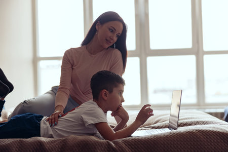 Side view on small teen boy lying on bed in bedroom and using laptop with mother.の写真素材