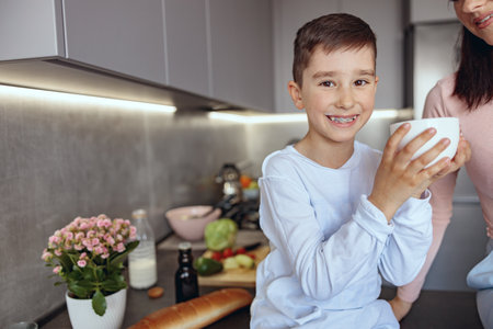 Portrait of cute little Caucasian boy smiling and holding big cup beside mom in kitchen.の写真素材