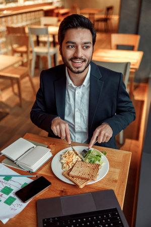 Indian businessman eating lunch during break after work in cafe and looking at sideの写真素材
