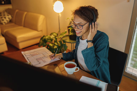 Smiling Afro american woman interior designer in headphones works with documents in home officeの写真素材