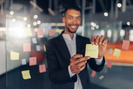 African Businessman showing sticky notes with motivational phrases while standing in officeの写真素材