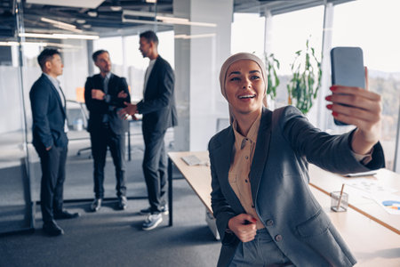 Smiling muslim business woman in hijab taking a selfie on colleagues background in officeの写真素材