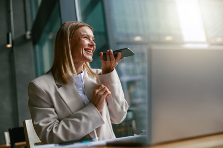 Happy businesswoman recording audio message on phone while while sitting in cozy cafe near windowsの写真素材