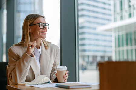 Smiling business lady holding pencil and drinking coffee while working in modern cafeの写真素材