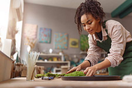Smiling Woman decorator making green ikebana on tray in florist workshop. Blurred backgroundの写真素材