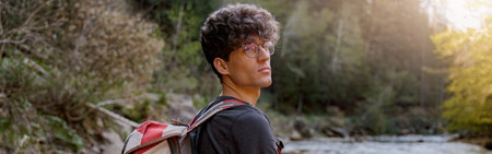 Handsome young male hiker with backpack standing at river in mountains. Trekking concept.