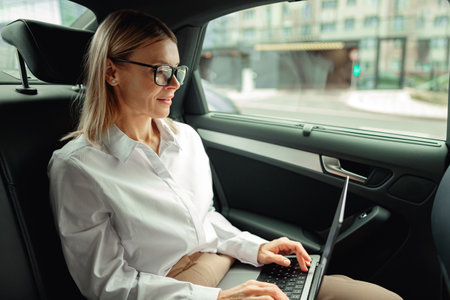Focused businesswoman in glasses working on laptop sitting on car backseat on the way to officeの写真素材