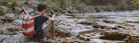 Rear on male tourist resting on rocks at river and using smartphone. Relax during trekking.