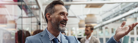 Mature smiling businessman standing in office on colleagues background and waving hi