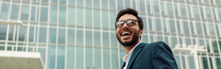 Smiling indian businessman in suit and glasses with laptop near office buildingの写真素材