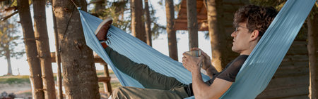 Handsome man having relax day in forest house. Sipping coffee in hammock in wood in morning.の写真素材