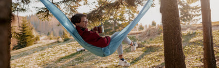 Cheerful woman in glasses resting in hammock i forest and drinking coffee from flask.