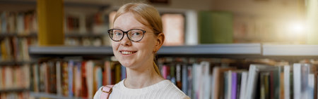 Female student in glasses chooses book while standing in the university library and looking camera