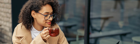 Afro american woman freelancer working remotely and drinking tea while sitting on cafe terrace