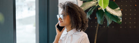 African businesswoman talking phone and holding laptop while standing near window in coworking