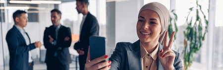 Smiling muslim business woman in hijab taking a selfie on colleagues background in office