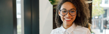 African businesswoman working laptop while standing near window in coworking and looking camera