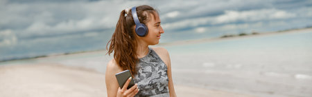 Smiling woman in sportswear listening music on headphones by the beach on background of oceanの写真素材