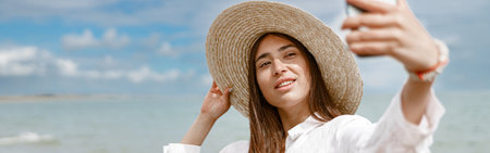 Beautiful woman in hat standing on beach near sea and making selphie