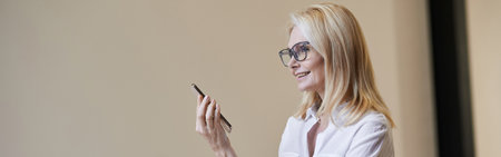 Busy hours. Stylish mature blonde woman in glasses and business attire smiling and talking on the...