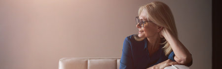 Happy mature woman in eyewear smiling while relaxing on a couch at home in the living roomの写真素材