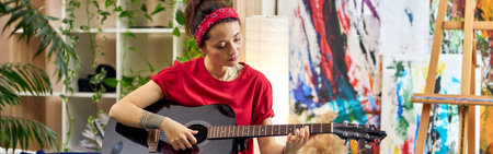Creative young woman playing guitar while sitting on a bed in modern studio apartmentの写真素材