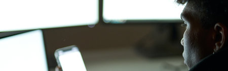 Close up shot of busy young guy sitting in dark room in front of many computer monitors and using...