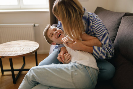 Overjoyed mother having fun with son at home, tickling and playing game on cozy couch in living roomの写真素材