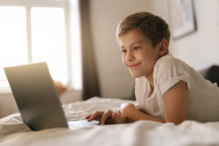 Young boy lies on the bed and looks into an open laptop. Home education conceptの写真素材