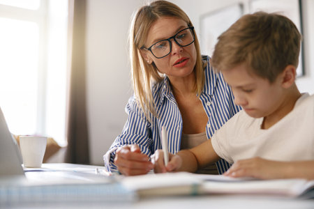 Schoolboy sitting at table and writing in copybook on online lesson while his mom helping him