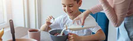 Small smiled son sitting at kitchen table with bowl of cereals, mother pouring milk.の写真素材