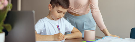 Caucasian little teen boy writing homework in copybook and mother standing by. Checking.の写真素材