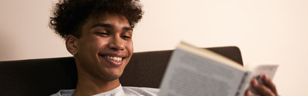 Amazing story. Portrait of cheerful brunette guy lying in the bed at home, smiling while reading interesting book at late eveningの写真素材