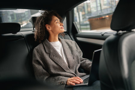 Woman freelancer working on laptop sitting on car backseat on way to office and looking at windowの写真素材
