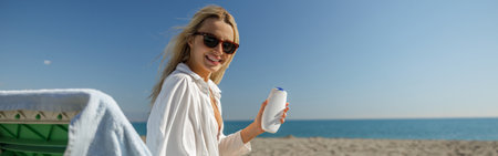 Blonde lady in sunglasses smiling at camera, advertising cream body lotion while tanning on a beach