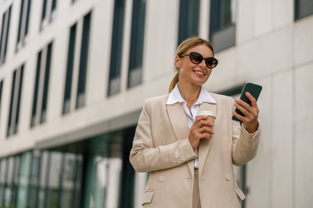 Woman manager use phone whiile standing on modern building background and drinking coffeeの写真素材