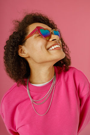 Woman in heart-shaped glasses with curly hair looking at side standing on pink studio background