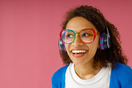 Smiling woman in headphones and glasses looking at side while standing on pink studio background