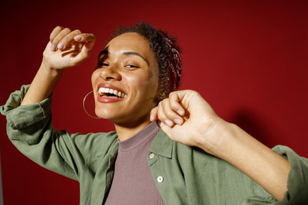 Close up of charming smiling woman looks away while dancing with raised hands on red background