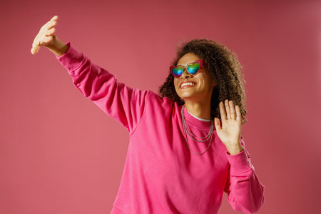 Woman in heart-shaped glasses raised hand closing from the light standing on pink studio background